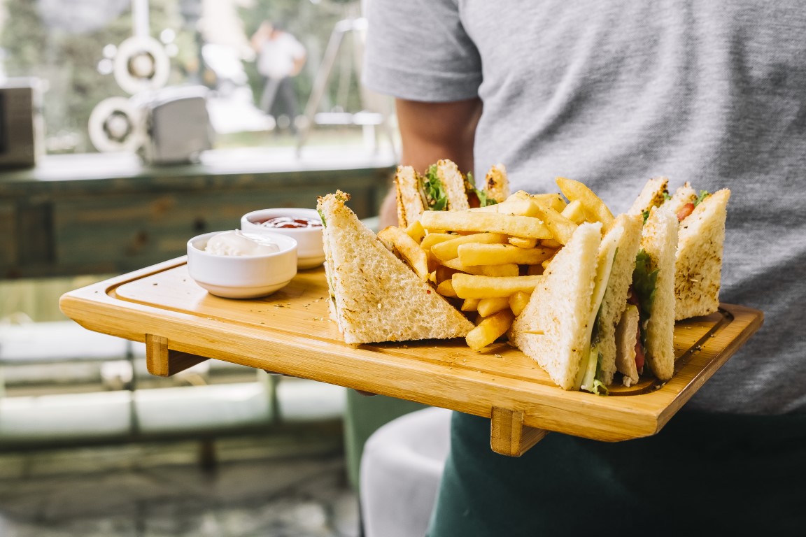 Man holding wooden board with club sandwich, bread toaster, chicken, tomato, cucumber, fries, mayonnaise, and ketchup on side view