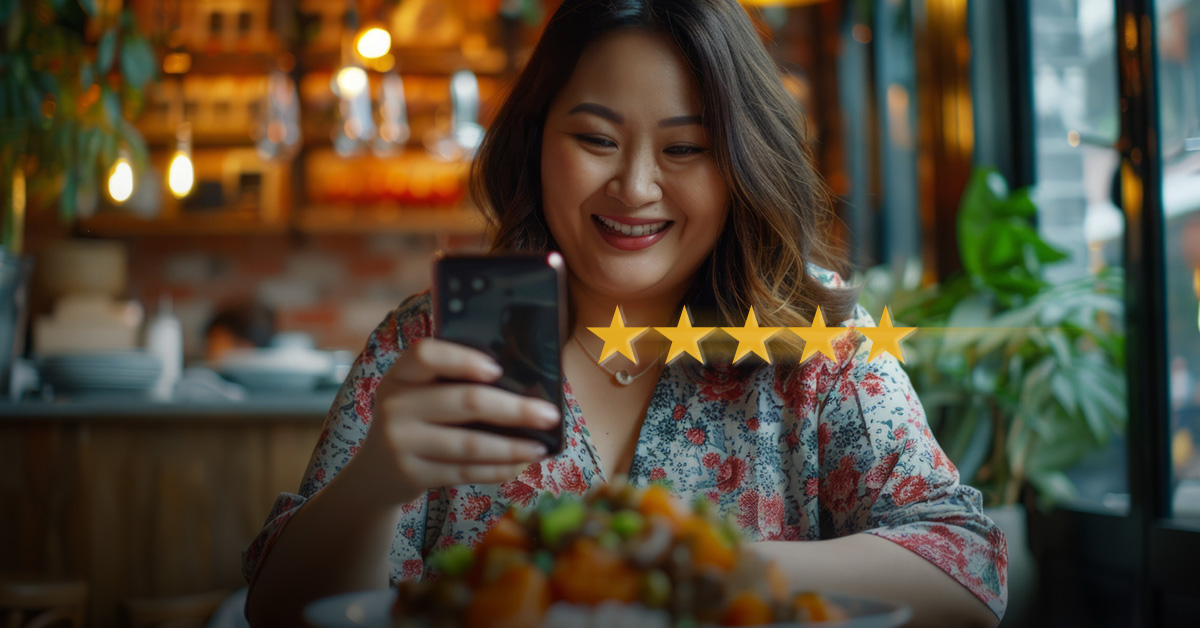 A woman smiles with a smartphone in her hand in a restaurant. She is giving a positive review.